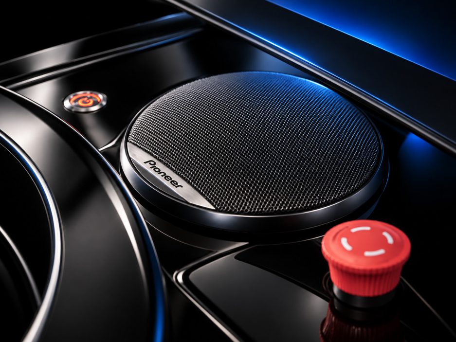 Imagem em close-up de um alto-falante automotivo Pioneer com grade de malha texturizada, embutido em um painel preto brilhante, iluminado por luz azul, com um botão de controle vermelho próximo.