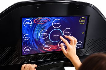 O persoană atinge un panou de control cu ecran tactil de pe un echipament de exerciții, selectând opțiunea 'Manual' dintre opțiunile circulare, cum ar fi Aerobic și Mers ușor.